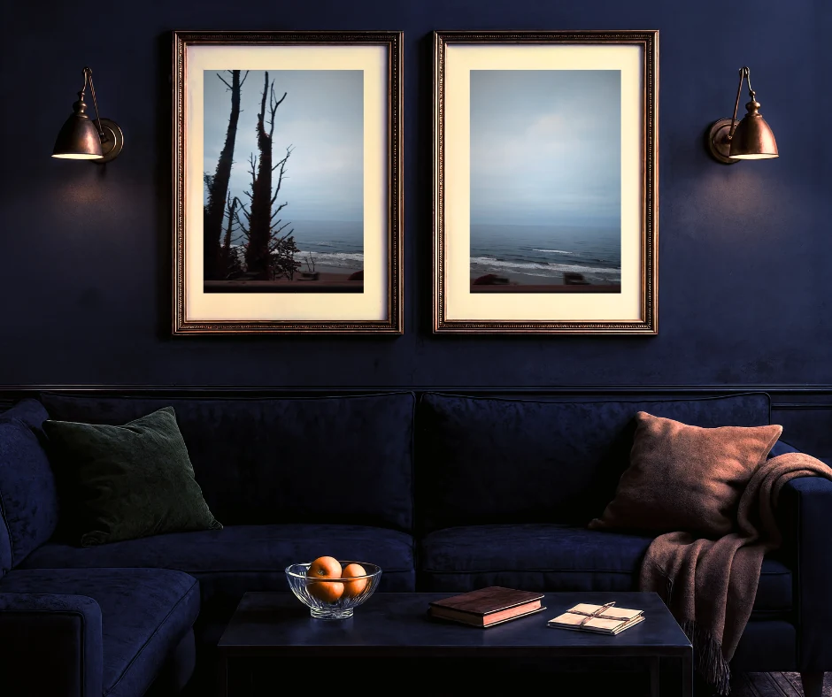 Deep-blue sitting room with two framed coastal photographs above a velvet sofa and journals on a low table