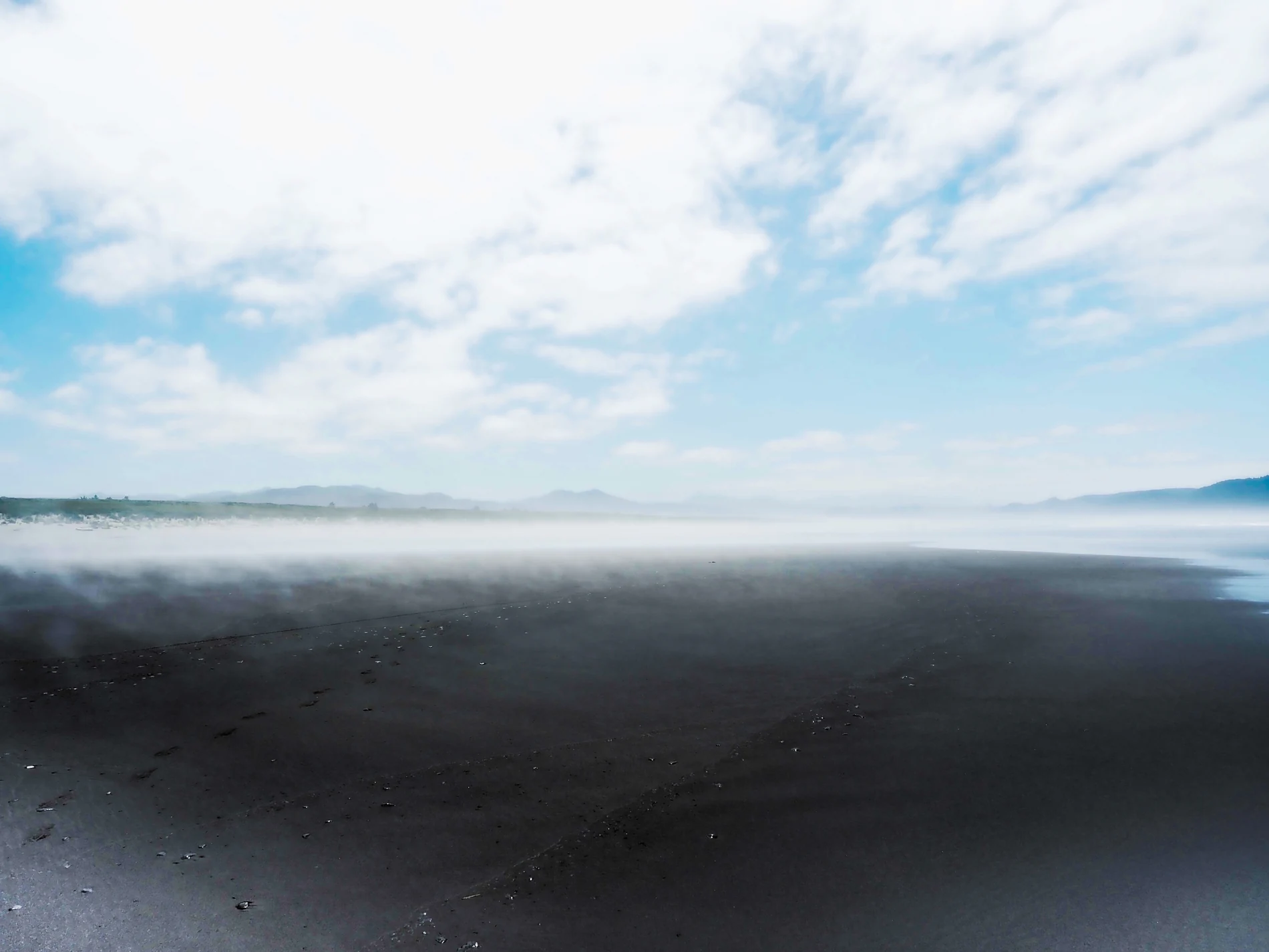 Mist drifting above a black-sand coast beneath a blue sky